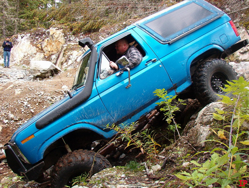 Blue 1985 Ford Bronco off-roading on a rocky trail, tilted on its side.