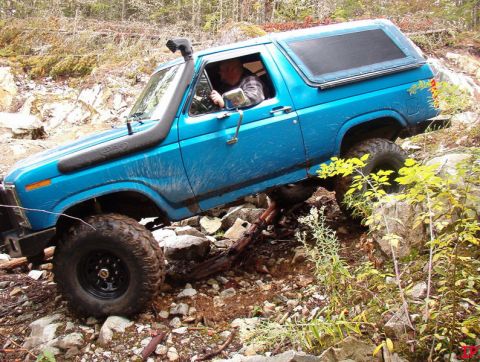 Blue 1985 Ford Bronco off-roading on a rocky trail with a snorkel.