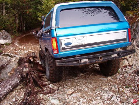 Teal Ford Bronco on a rocky, wooded trail, tilted right.