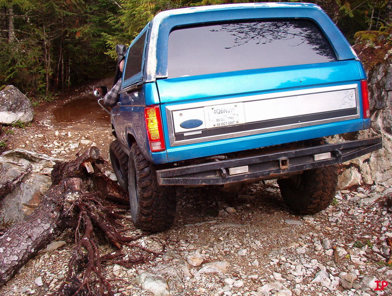 Teal Ford Bronco on a rocky, wooded trail, tilted right.
