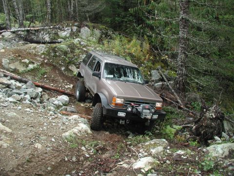 Tan SUV with off-road modifications on a rocky, wooded trail.
