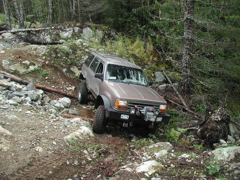 Tan SUV with off-road modifications on a rocky, wooded trail.