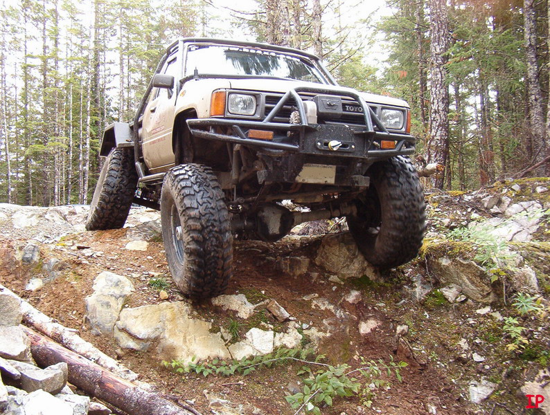 Modified tan Toyota pickup truck with large off-road tires on a rocky, wooded trail.