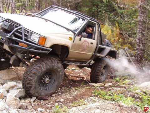 Tan Toyota pickup truck with large off-road tires on a rocky, wooded trail, driver visible.