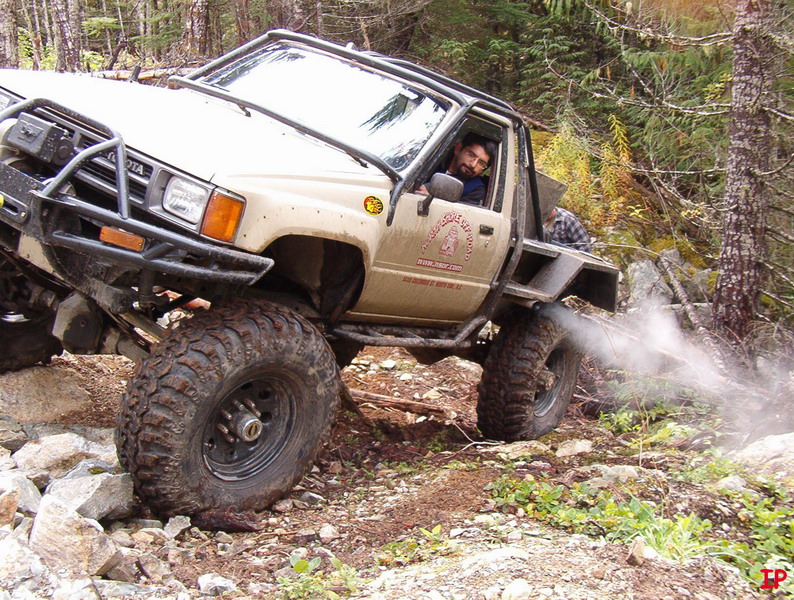 Tan Toyota pickup truck with large off-road tires on a rocky, wooded trail, driver visible.