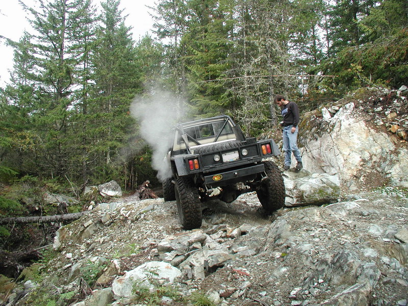 Off-road truck on a rocky trail, emitting smoke, with a person watching.