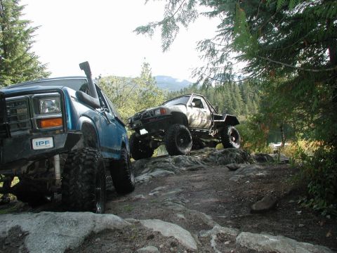 Two off-road trucks on a rocky trail with trees and mountains in the background.