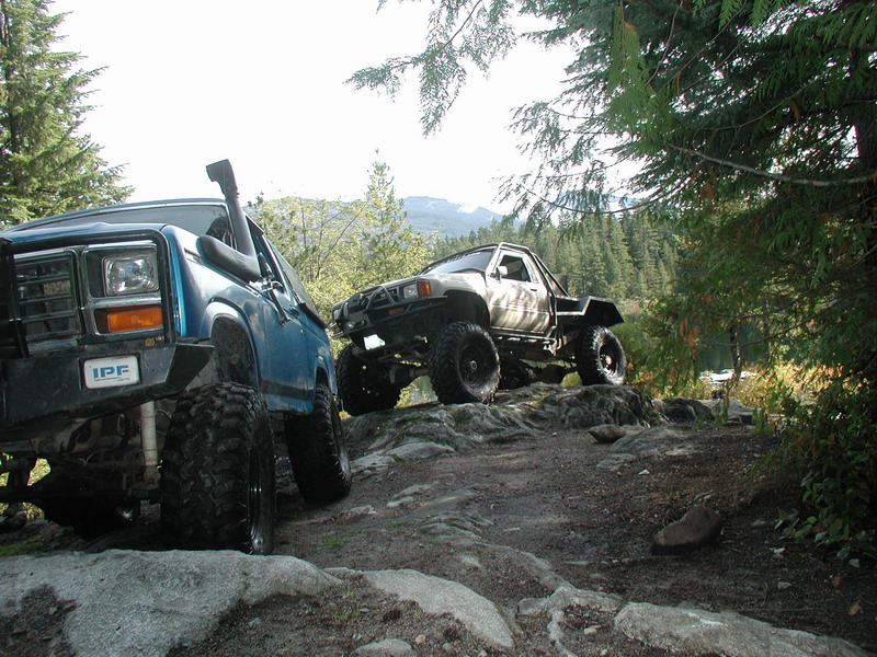 Two off-road trucks on a rocky trail with trees and mountains in the background.
