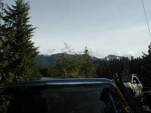 Rear of a blue SUV with a snorkel, parked near a white off-road vehicle in a forest with mountains.