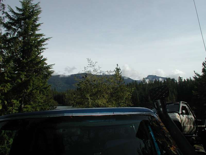 Rear of a blue SUV with a snorkel, parked near a white off-road vehicle in a forest with mountains.