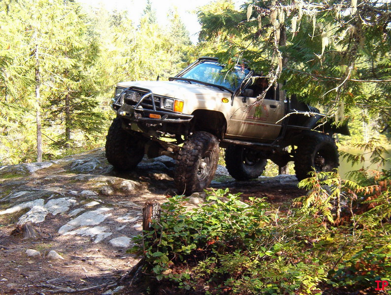 Lifted tan pickup truck on rocky terrain with trees and water.