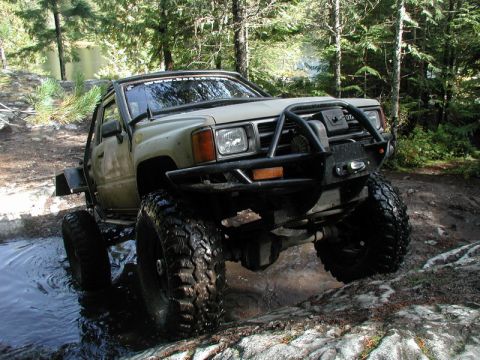 Lifted olive green Toyota 4x4 truck with large tires driving through water and rocks in a forest.
