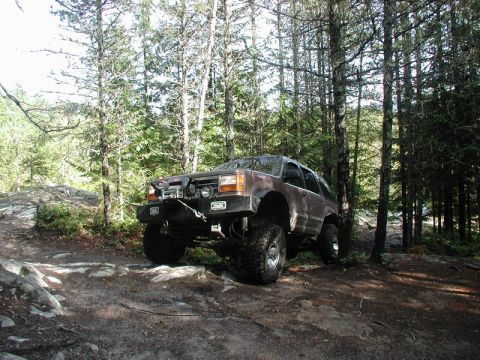 Lifted brown SUV with winch on a rocky, wooded trail.