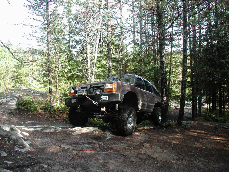 Lifted brown SUV with winch on a rocky, wooded trail.