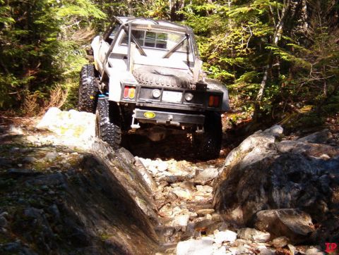 Off-road pickup truck with large tires and roll cage driving on a rocky forest trail.