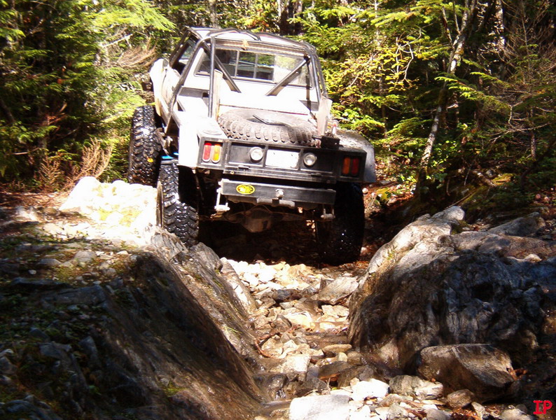 Off-road pickup truck with large tires and roll cage driving on a rocky forest trail.