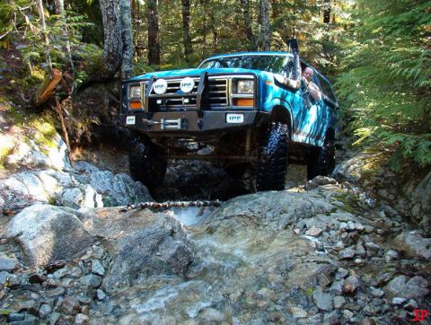 Blue Ford Bronco off-roading through a rocky stream in a forest.