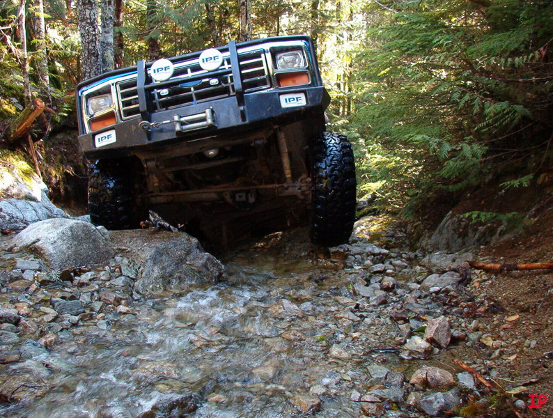 1985 Ford Bronco navigating a rocky stream in a forest.