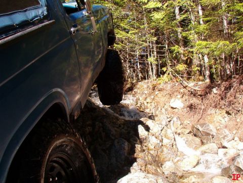 Teal 1985 Ford Bronco driving over rocks and water in a forest.