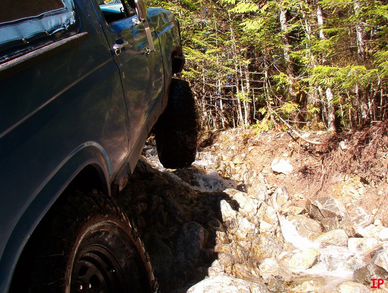 Teal 1985 Ford Bronco driving over rocks and water in a forest.