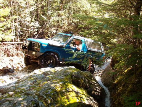 Blue 1985 Ford Bronco off-roading through a rocky stream in a forest.
