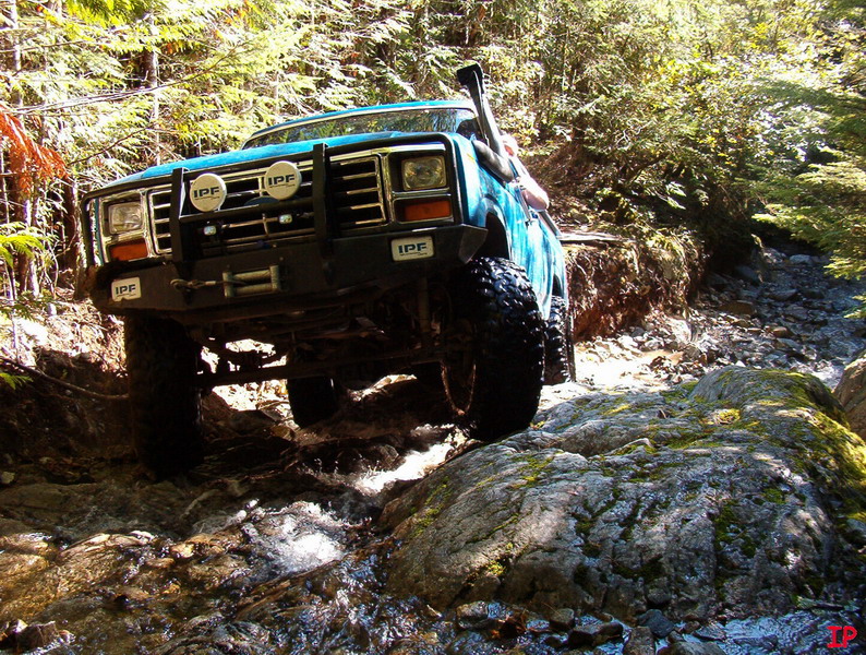 Blue 1980s Ford Bronco off-roading through a rocky stream.