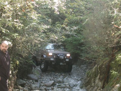 Brown SUV driving through a rocky stream in a forest.