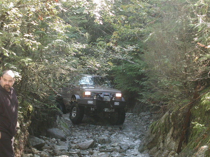 Brown SUV driving through a rocky stream in a forest.