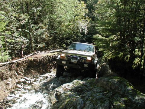 Tan Jeep Cherokee XJ off-roading through a rocky stream in a forest.