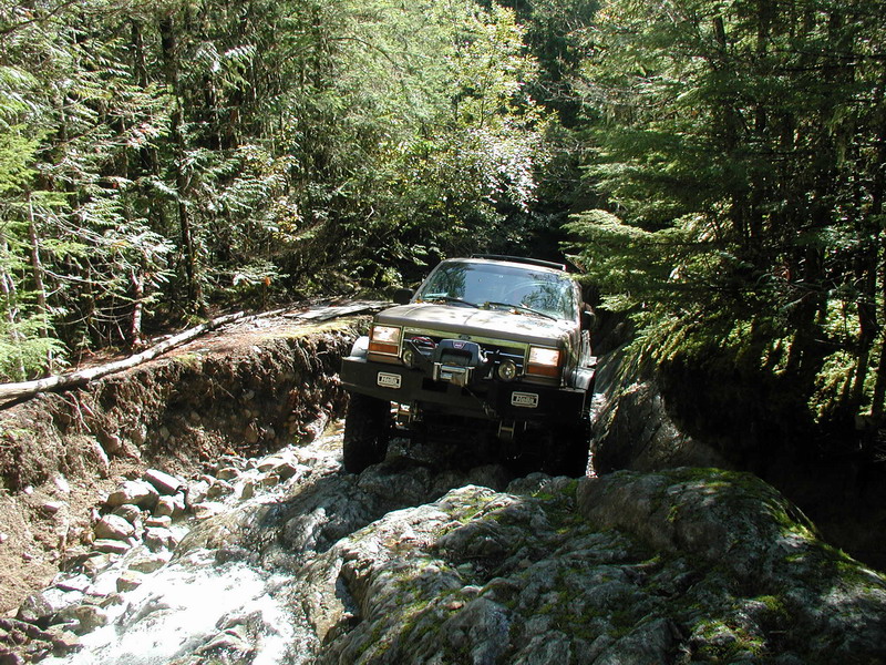 Tan Jeep Cherokee XJ off-roading through a rocky stream in a forest.