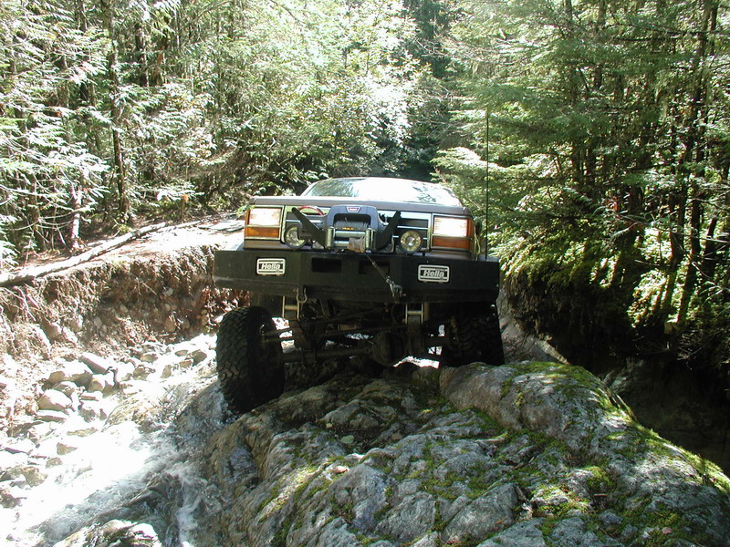 Modified dark brown SUV driving through a rocky stream in a forest.