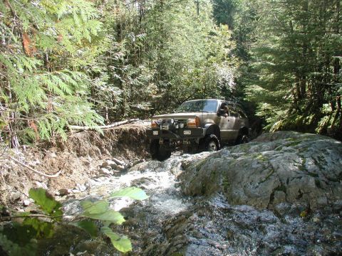 Brown SUV off-roading through a rocky stream in a forest.