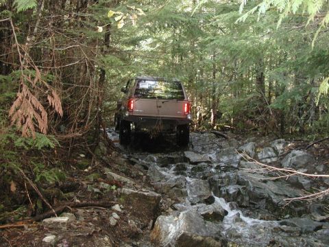Brown SUV driving through a rocky stream in a forest.