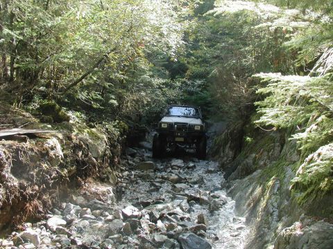 White Jeep Cherokee driving through a rocky stream in a forest.