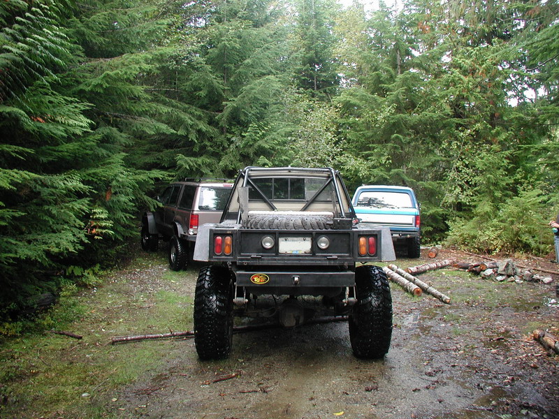 Rear view of a custom flatbed truck with large tires, parked in a forest with other vehicles.