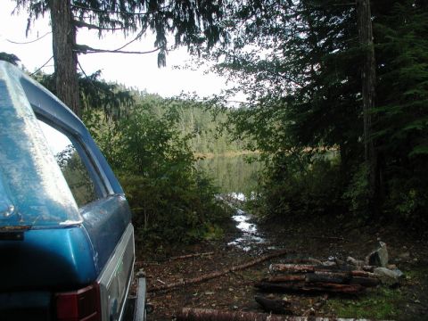 Blue Ford Bronco parked by a lake with a campfire pit in the woods