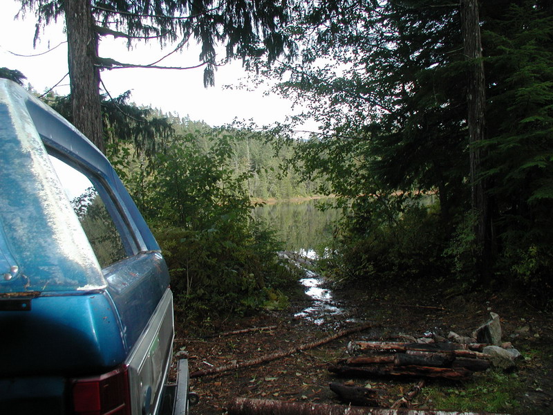 Blue Ford Bronco parked by a lake with a campfire pit in the woods