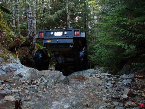 Rear view of a flatbed truck driving through a rocky stream in a forest.