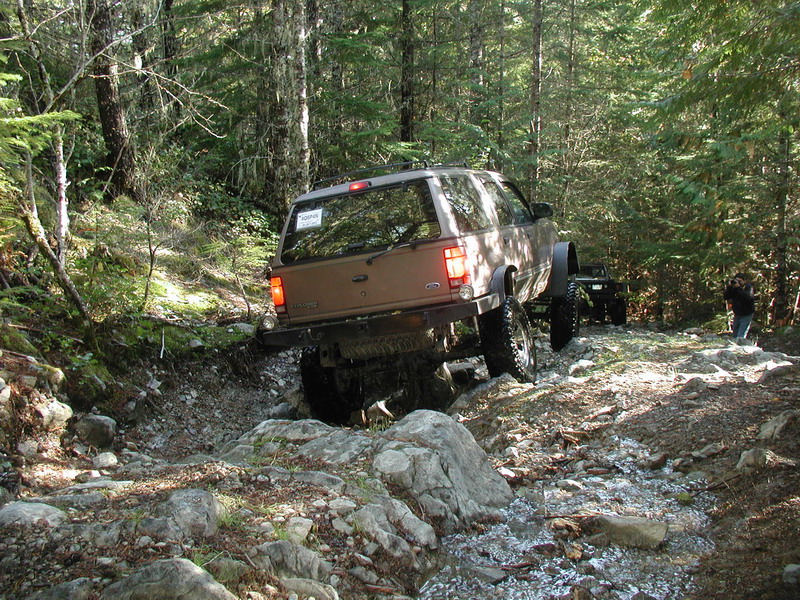Lifted brown Ford Explorer driving on a rocky trail in a forest.