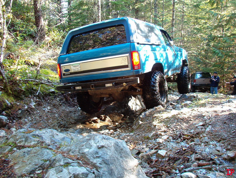 Teal Ford Bronco off-roading on a rocky trail in a forest.
