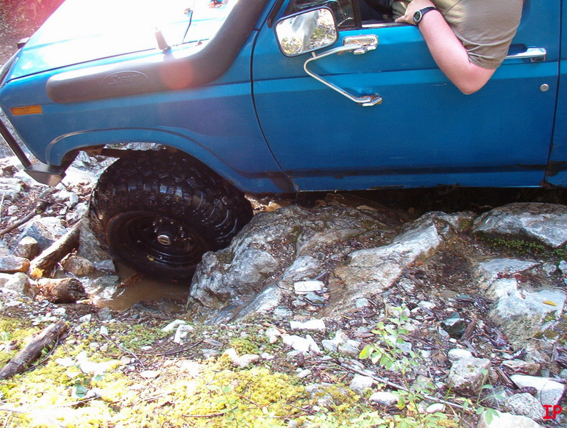 Blue Ford Bronco off-roading over rocks, front passenger tire stuck in mud and rocks.