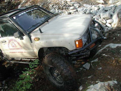 Beige off-road SUV on rocky terrain with custom roll cage and large tires.