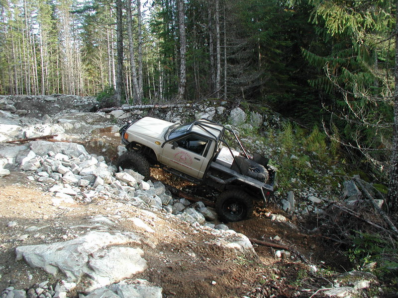 Tan Toyota pickup truck with roll cage on a rocky, wooded trail.