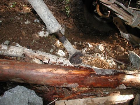 Damaged driveshaft on logs with vehicle tire and suspension visible.