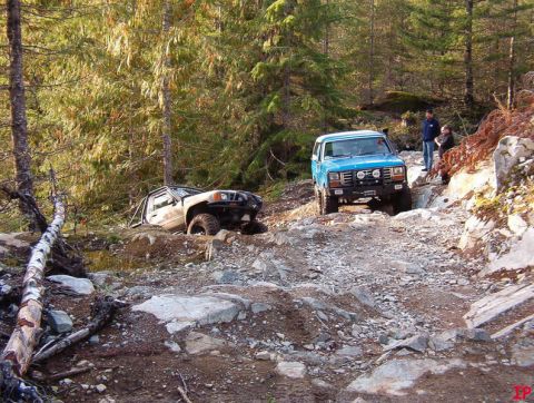 Blue 1985 Ford Bronco driving on a rocky forest trail, another vehicle stuck on the side.