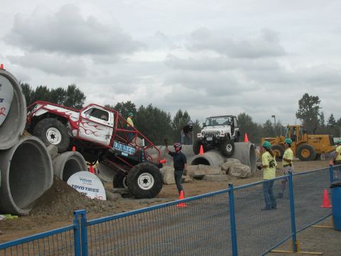 Custom monster truck with red flames on concrete pipes, Jeep in background, people watching.
