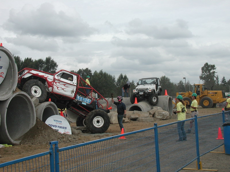 Custom monster truck with red flames on concrete pipes, Jeep in background, people watching.