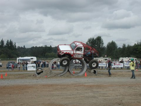Custom off-road truck with red roll cage driving over concrete pipes with spectators in background.