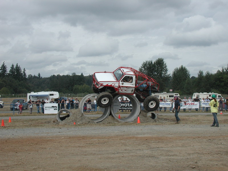 Custom off-road truck with red roll cage driving over concrete pipes with spectators in background.