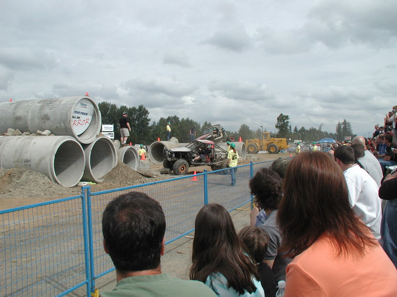 Custom off-road buggy airborne over dirt and concrete pipes at an event with spectators.